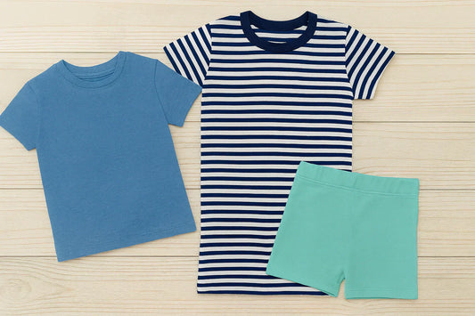lat lay of budget-friendly girlish shirts and baby clothes for boys featuring a solid blue T-shirt, a striped navy and white T-shirt, and mint green shorts on a wooden background.